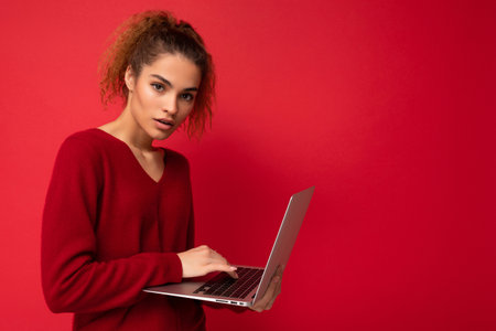 Close-up portrait of beautiful dark blond woman holding laptop computer looking at camera typing on keyboard wearing red sweater isolated over red wall background.の写真素材