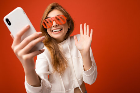 Attractive charming young smiling happy woman holding and using mobile phone taking selfie wearing...