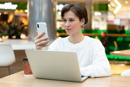 girl in a cafe sits at a laptop, looks at the phone screen. Brunette freelancer in white sweater.