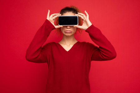 happy woman blinks eyes, with blank screen, shows modern device, isolated over red background. Technology concept.の写真素材