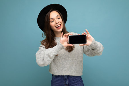 Attractive young smiling woman wearing black hat and grey sweater holding smartphone looking down...