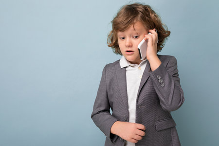 Photo of Handsome boy with curly hair wearing grey suit holding and using phone isolated over blue background looking downand talking on mobile with somebody. Copy spaceの写真素材