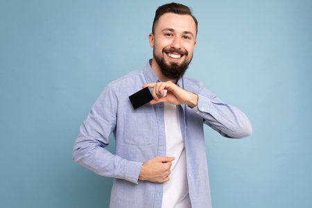 Good looking cool smiling brunette bearded young man wearing stylish blue shirt and white t-shirt isolated over blue background wall holding credit card looking at camera.の写真素材