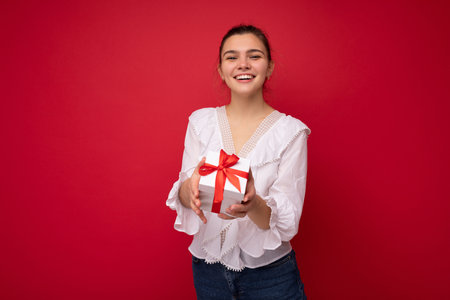 Beautiful happy young brunette woman isolated over colourful background wall wearing stylish casual clothes holding gift box and looking at camera. Empty spaceの写真素材