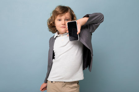 Handsome positive boy with curly hair wearing suit holding phone isolated over blue background...