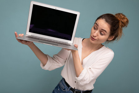 Beautiful charming young woman holding netbook computer looking down wearing white shirt isolated on blue background. Mock upの写真素材