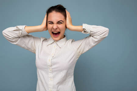 Portrait of emotional young beautiful brunette woman with sincere emotions wearing white shirt isolated over blue background with copy space and covering ears with hands and yelling.の写真素材