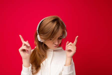 Attractive emotional positive young blonde woman wearing white hoodie isolated over colourful background wearing white wireless bluetooth headsets listening to music and dancing.の写真素材