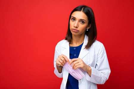 Woman holding virus protective mask on face against  and white medical coat isolated on red...