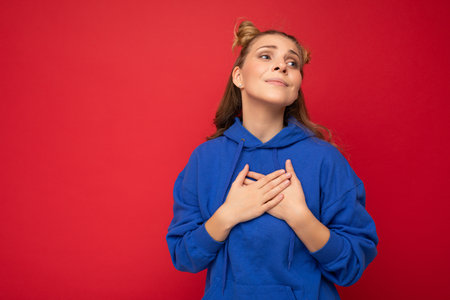 Portrait of young happy positive pretty cute blonde woman with two horns with sincere emotions wearing stylish clothes isolated on red background with free space and holding hands on chest.の写真素材