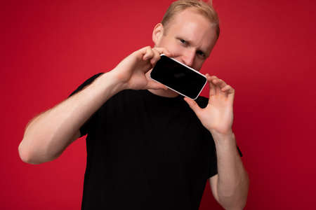 Amazing handsome blonde young man wearing black t-shirt standing isolated on red background with...