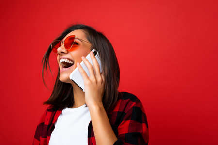Closeup photo of attractive positive laughing young brunet woman wearing stylish red shirt white t-shirt and red sunglasses isolated over red background communicating on mobile phone looking up.の写真素材
