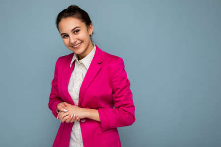 Portrait of young attractive beautiful positive smiling brunette woman with sincere emotions wearing casual white shirt and trendy pink jacket isolated over blue background with free space.の写真素材