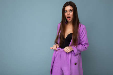 Portrait of shocked amazed beautiful fashionable brunette woman in casual trendy violet jacket isolated on blue background with free space. Surprise concept.の写真素材