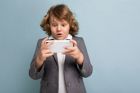 Photo shot of Handsome amazed emotional child boy with curly hair wearing grey suit holding and using phone isolated over blue background looking at smartphone playing games.の写真素材
