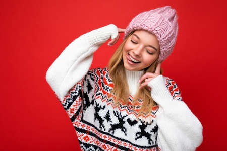 Photo of beautiful happy funny joyful young blonde woman isolated over red background wall wearing winter sweater and pink hat looking down and having fun.の写真素材