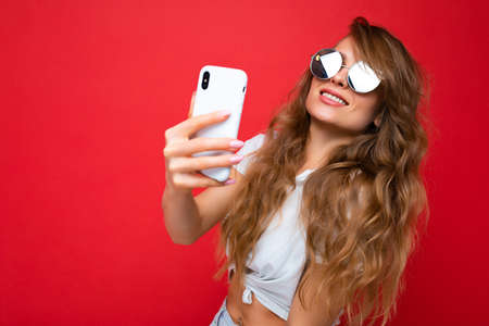 Attractive charming young smiling happy woman holding and using mobile phone taking selfie wearing stylish clothes isolated over wall background.の写真素材