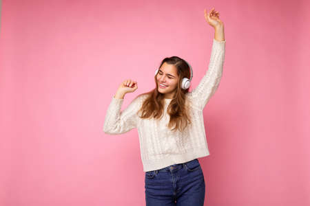 Attractive positive smiling young brunette curly woman wearing white sweater isolated on pink...