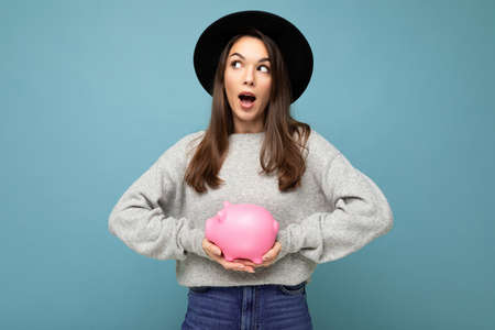 Portrait of shocked amazed young beautiful brunette woman wearing stylish gray sweater and black hat isolated over blue background with free space and holding pink piggy box. Money box concept.の写真素材