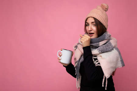Beautiful sick young brunet woman wearing black sweater hat and warm scarf isolated over pink background holding paper mug for mockup drinking hot tea and looking at camera and caughing. copy spaceの写真素材