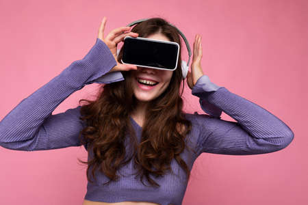 Photo of pretty positive smiling young brunette curly woman wearing purple crop top isolated over pink background wall holding and suggesting mobile phone with empty display and bluetooth headsets.の写真素材