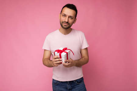 Photo shot of handsome positive brunette unshaven young man isolated over pink background wall wearing pink t-shirt holding white gift box with red ribbon and looking at camera.の写真素材