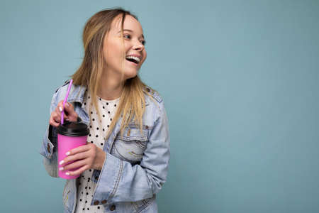 Charming young happy blonde woman wearing stylish clothes isolated over colourful background wall holding paper cup for mockup drinking milkshake looking to the side. empty spaceの写真素材