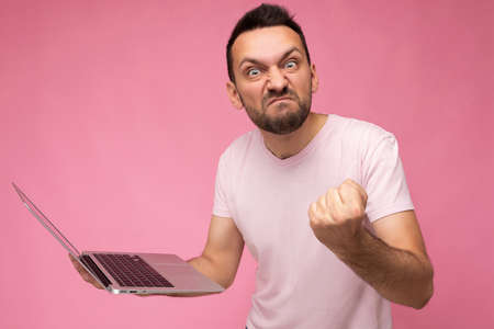 Handsome angry brunet man holding laptop computer showing fist looking at camera in t-shirt on...