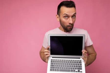 Handsome surprised and amazed brunet man holding laptop computer looking at camera in t-shirt on...