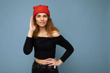 Photo of young positive happy beautiful brunette curly woman with sincere emotions wearing black crop top and red hat isolated on blue background with copy space and listening attentivelyの写真素材