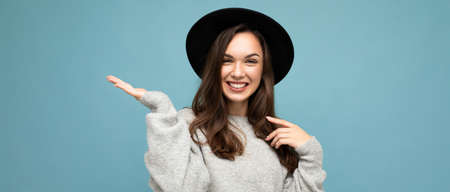 Panoramic photo of attractive brunette happy smiling young woman pointing hand at free space for text wearing grey jersey and black hat isolated on blue backgroundの写真素材