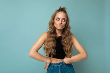 Shot of young thoughtful pretty blonde woman wearing black top isolated on blue background with empty space and having doubtsの写真素材