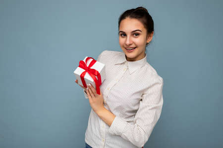 Beautiful happy young brunette woman isolated over colourful background wall wearing stylish casual clothes holding gift box and looking at cameraの写真素材