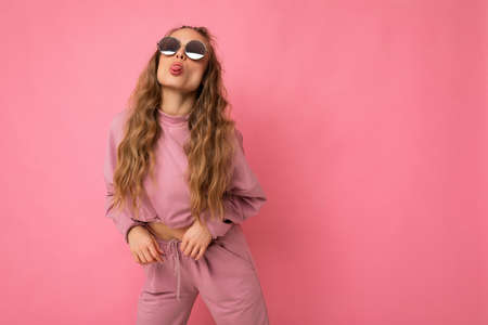 Shot of fascinating positive happy young dark blonde curly woman isolated over pink background wall wearing casual pink sport clothes and stylish sunglasses and having funの写真素材