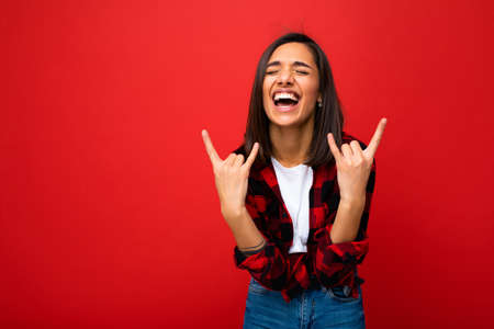 Photo of young happy positive cool attractive brunette woman with sincere emotions wearing white t-shirt and hipster red check shirt isolated on red background with empty space and showing rock signの写真素材
