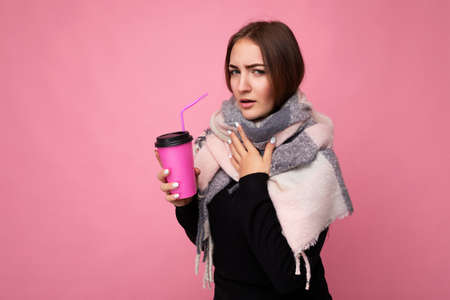Photo shot of beautiful young brunette woman wearing black sweater and warm scarf isolated over pink background holding paper coffee cup for mockup drinking hot beverage and having coldの写真素材