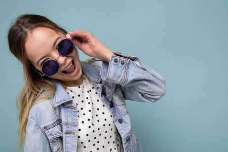 Photo shot of beautiful positive surprised young blonde woman wearing summer casual clothes and stylish sunglasses isolated over colorful background looking at cameraの写真素材
