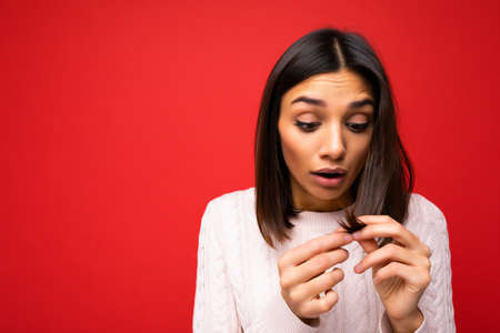 Portrait of beautiful shocked amazed young brunette woman wearing knitted jersey isolated over red background with free space and touching short hair and having split endsの写真素材