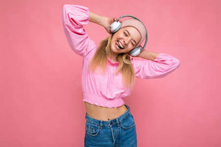 Photo of attractive positive smiling young blonde woman wearing pink blouse and pink hat isolated over pink background wall wearing white wireless bluetooth earphones listening to music having funの写真素材