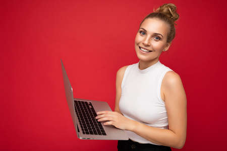 Side profile photo shot of beautiful smiling blond young woman with gathered hair wearing white t-shirt holding computer laptop typing on keyboard looking at camera isolated over red wall backgroundの写真素材