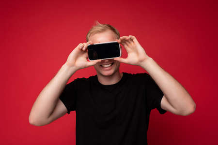 Portrait of smiling handsome blonde young man wearing black t-shirt standing isolated on red...