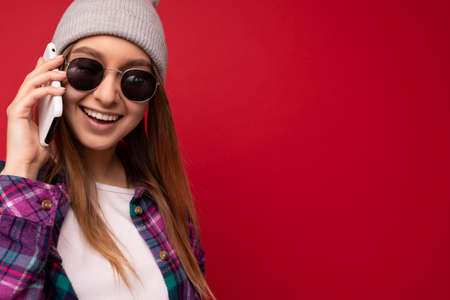 Closeup photo of beautiful happy positive young blonde woman wearing hipster purple shirt and casual white t-shirt grey hat and sunglasses isolated over red background holding in hand and talking on mobile phone looking at camera. Copy spaceの写真素材