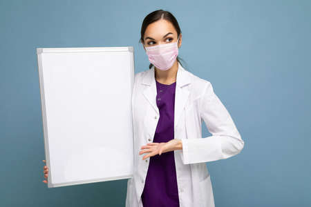 Beautiful brunette female nurse in protective face mask and white medical coat holding an empty...