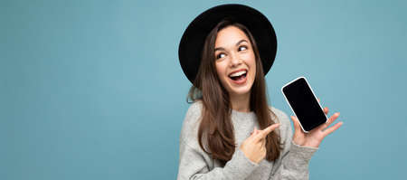 Panoramic Closeup of charming young happy woman wearing black hat and grey sweater holding phone...