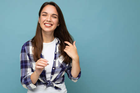 Young positive smiling beautiful brunette woman with sincere emotions wearing trendy check shirt standing isolated on blue background with empty spaceの写真素材