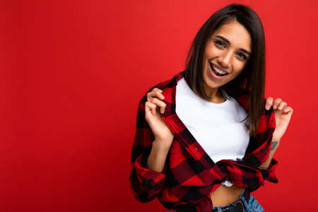 Young happy positive cool pretty brunette woman with sincere emotions wearing white t-shirt and trendy red check shirt isolated on red background with free spaceの写真素材