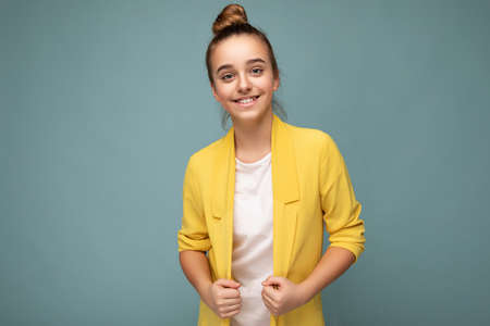 Shot of beautiful happy smiling brunette little girl wearing trendy yellow jacket and white t-shirt standing isolated over blue background wall looking at cameraの写真素材