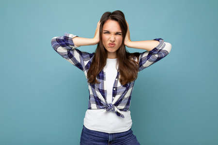 Dont want to hear it. Young emotional dissatisfied beautiful brunette woman wearing check shirt isolated on blue background with copy space and covering earsの写真素材