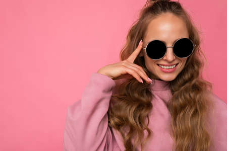 Closeup photo of pretty positive smiling young blonde curly woman isolated over pink background wall wearing casual pink sport clothes and stylish sunglasses looking at cameraの写真素材
