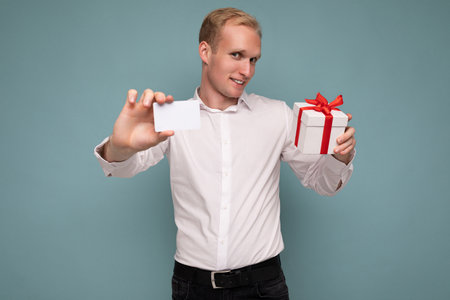 Handsome positive blonde man wearing white shirt isolated over blue background wall holding credit card and gift box looking at cameraの写真素材